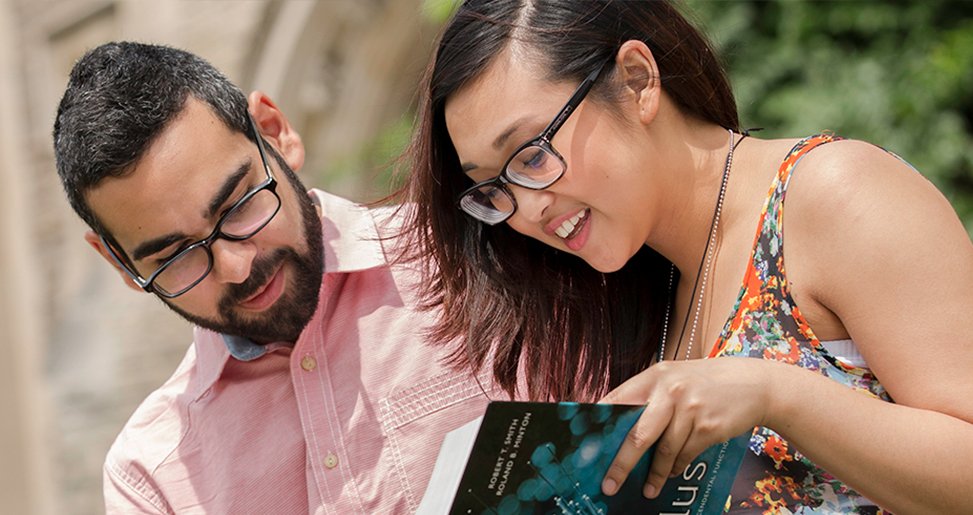 Two students standing outside looking at a textbook together.